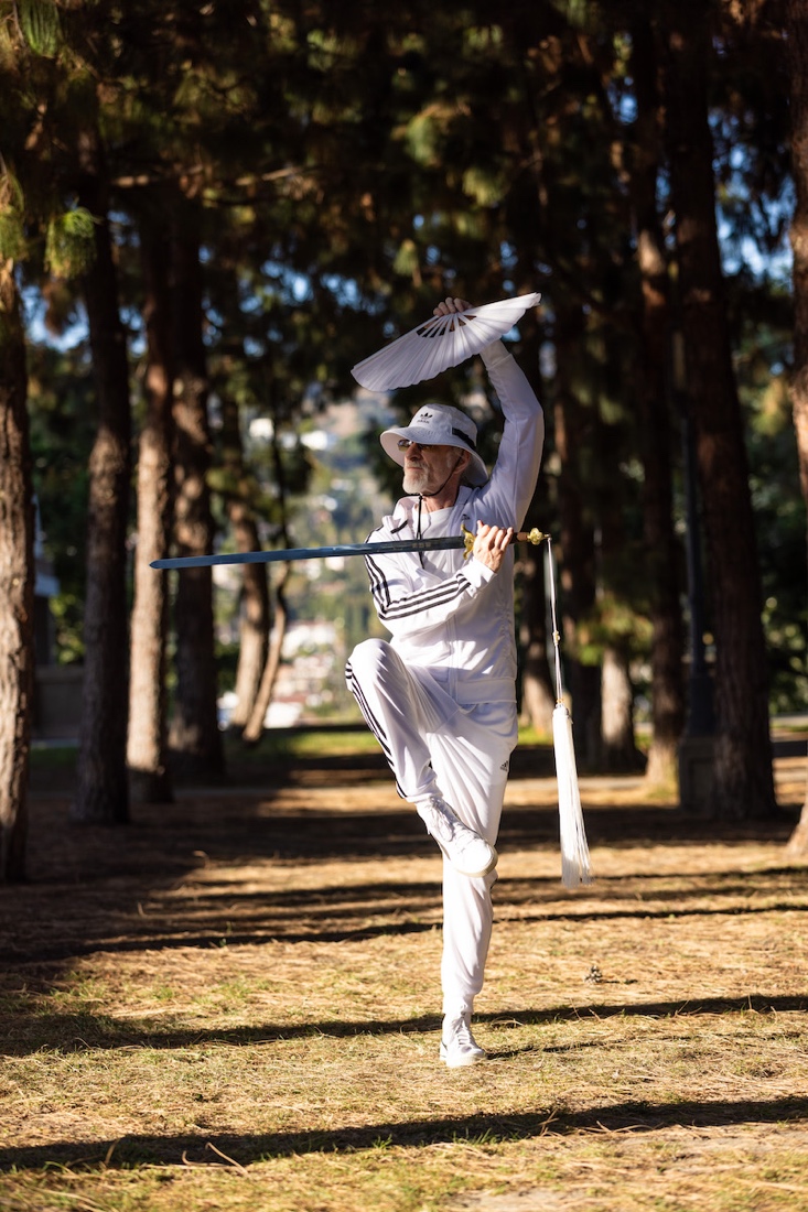 A person in white clothes holding a fan and standing on one leg
Description automatically generated
